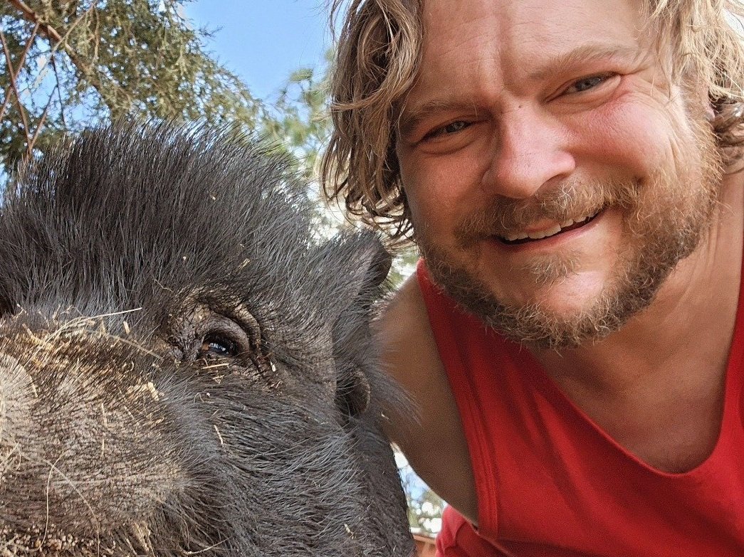 A man smiling for a selfie with a pig outdoors in a wooded area.